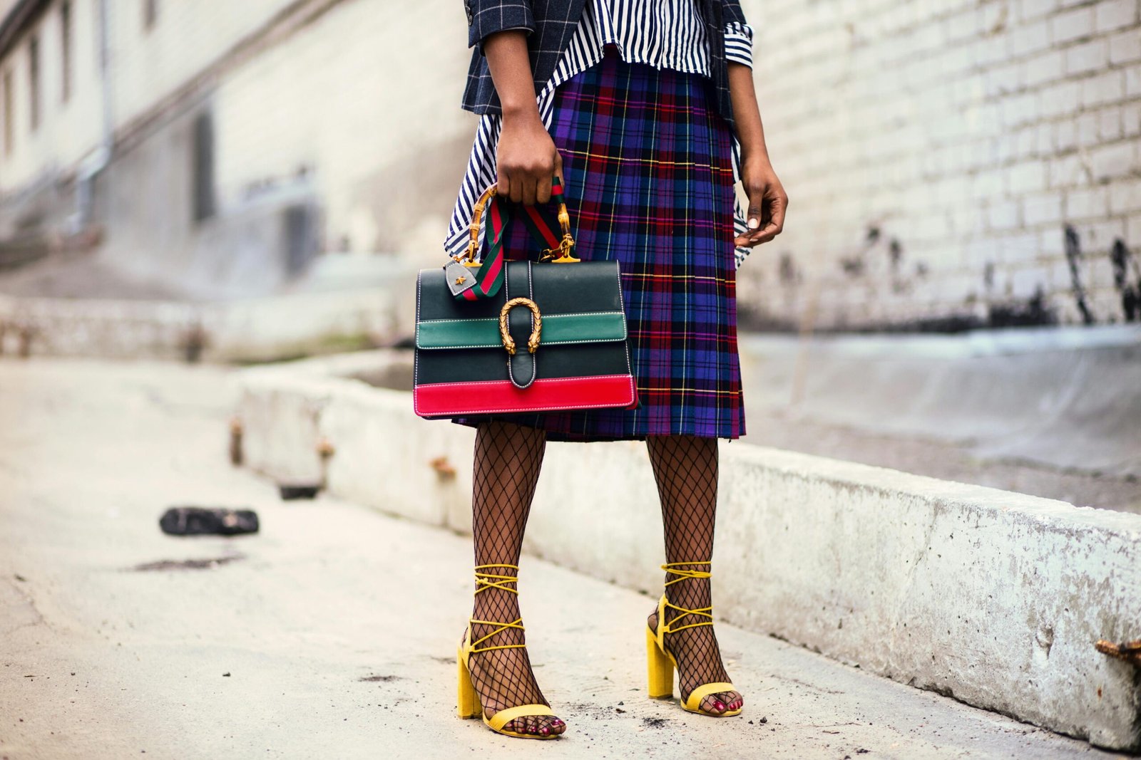 A woman wearing a bold personal style outfit with a plaid midi skirt, striped shirt, fishnet tights, yellow heels, and a statement handbag on a city street.