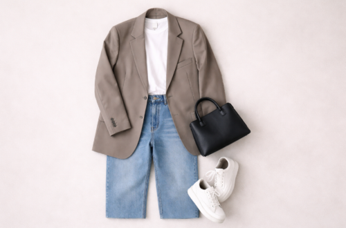 Model off duty style flat lay outfit featuring oversized neutral blazer, white T-shirt, straight-leg jeans, white sneakers and structured handbag on a minimal beige background.