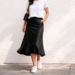 Black satin midi slip skirt styled with a white T-shirt and white sneakers in a minimalist everyday outfit, showing the fluid drape and bias-cut silhouette.